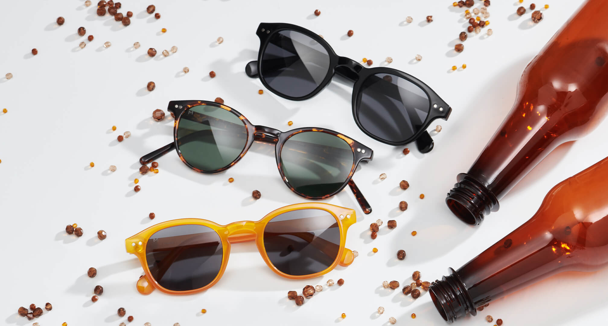 trīs saulesbriļļu pāri, kas izgatavoti no ilgtspējīgiem materiāliem uz balta galda, ar pērlēm un plastmasas pudelēm apkārt.